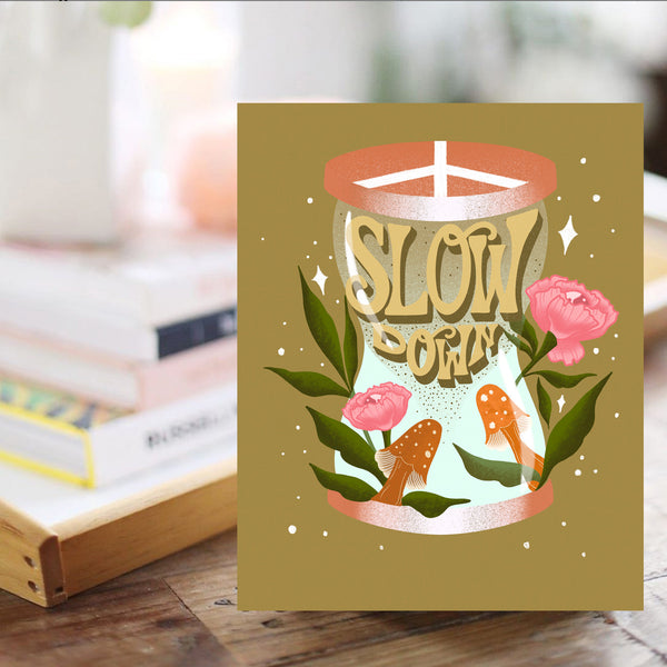 A groovy, retro inspired art print sits in front of a bright sunny scene with a pile of books and plants. A framed art print - the illustration is of an hour glass with the hand lettered message "Slow Down" inside, falling apart like sand. It is surrounded by vibrant orange mushrooms and pink flowers and greenery. The lid of the hour glass is a peace sign.