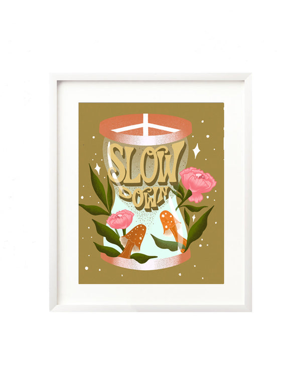A framed art print - the illustration is of an hour glass with the hand lettered message "Slow Down" inside, falling apart like sand. It is surrounded by vibrant orange mushrooms and pink flowers and greenery. The lid of the hour glass is a peace sign.