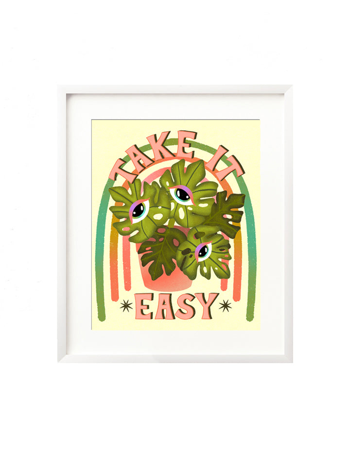 A framed art print - the illustration is of a monstera plant with colorful intuitive eyes in each leaf. It is in a vibrant pink pot, with a colorful green, teal, pink, and yellow rainbow behind it. Hand lettering spells out "Take it Easy" in a bold, groovy, retro inspired style.